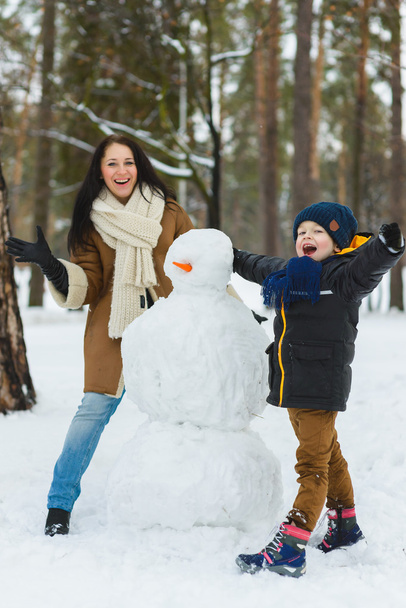 Happy family in warm clothing. Smiling mother and son making a snowman outdoor. The concept of winter activities - Photo, Image