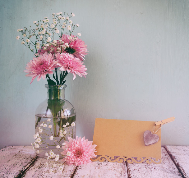 fresh white and pink flowers, heart next to vintage empty card over wooden table. vintage filtered and toned image - Фото, зображення
