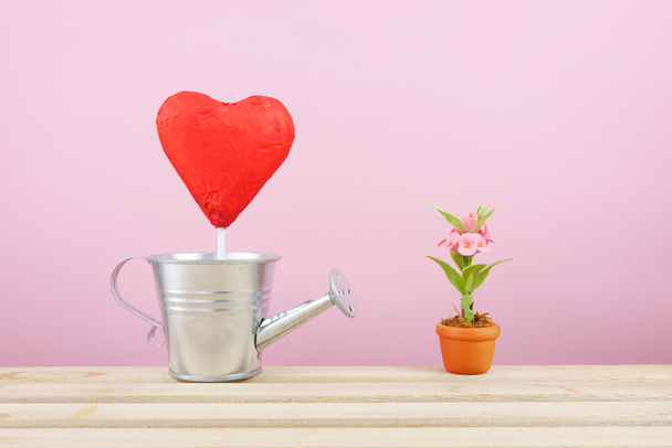 El palo de corazón de chocolate frustrado rojo con una pequeña regadera de plata y mini flor falsa en maceta de planta marrón en bandeja de madera
 - Foto, Imagen