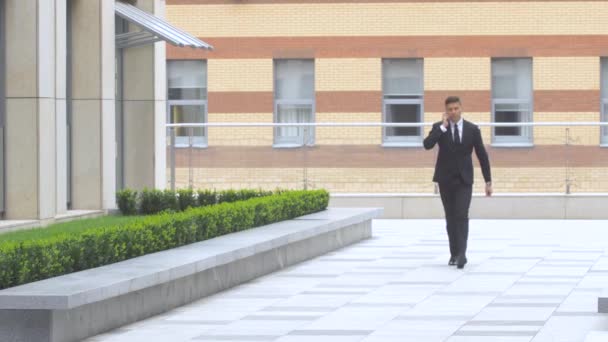 businessman speaking on the phone while walking along modern building - Materiał filmowy, wideo
