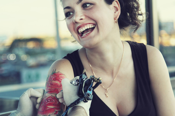 tattooer δείχνει τη διαδικασία της λήψης ένα τατουάζ για νέους hipster όμορφη γυναίκα με τα κόκκινα μαλλιά σγουρά. Σχέδιο δερματοστιξιών υπό μορφήν τριαντάφυλλο - Φωτογραφία, εικόνα