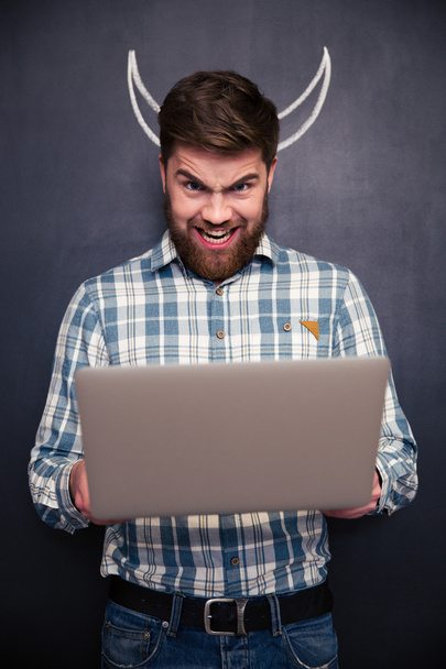 Funny man using laptop over blackboard background with drawn horns - Photo, Image