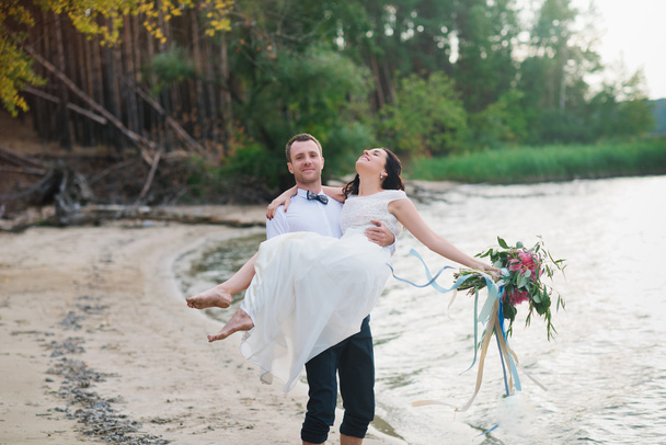 Jovem noivo bonito segura sua noiva em seus braços na praia com um grande buquê de flores bonitas
 - Foto, Imagem