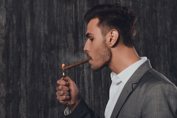 Handome hombre brutal en traje sobre el fondo gris iluminación de un cigarro
 - Foto, Imagen