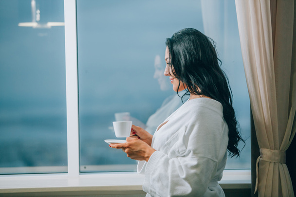 Getting warm with fresh coffee. Beautiful young woman in white bathrobe drinking coffee and looking through a window - Photo, image