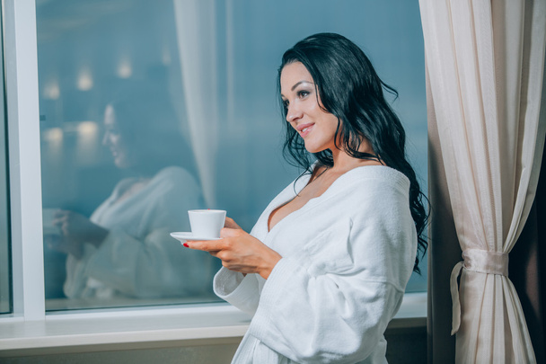 Steeds warm met verse koffie. Mooie jonge vrouw in witte badjas koffie drinken en door een venster kijken - Foto, afbeelding
