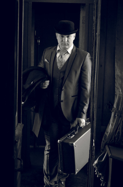 Монохромное фото человека в шляпе с чемоданом, идущего в трамвае
 - Фото, изображение
