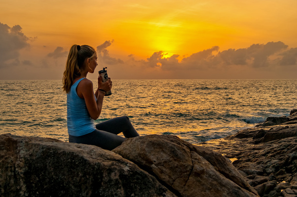 Woman sitting at dawn on the cliff near a sea,holding bottle and watching sunrise on a tropical island - Foto, afbeelding