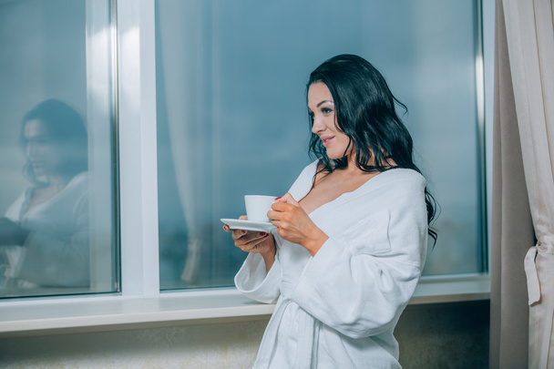 Getting warm with fresh coffee. Beautiful young woman in white bathrobe drinking coffee and looking through a window - Zdjęcie, obraz