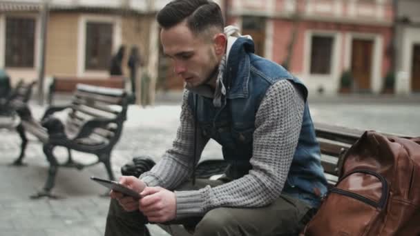 Przystojny młody człowiek siedzi na ławce w centrum miasta Europejskiego za pomocą tabletu. Facet gra online gry na swoim urządzeniu mobilnym. - Materiał filmowy, wideo