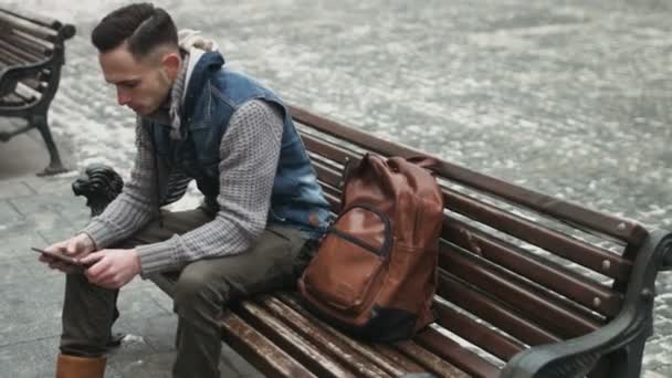 . Fiatal férfi használja a tablettát a szabadban, majd tedd be a barna hátizsák. Guy ül a padon belül egy régi város, és megpróbálta elrejteni a mobil eszközt a zsákba. - Felvétel, videó