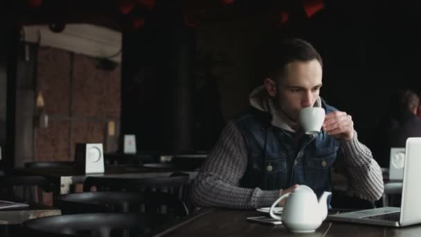 Hombre joven trabajando en el ordenador portátil dentro de un acogedor café medio vacío y haciendo un descanso para tomar té caliente
. - Imágenes, Vídeo
