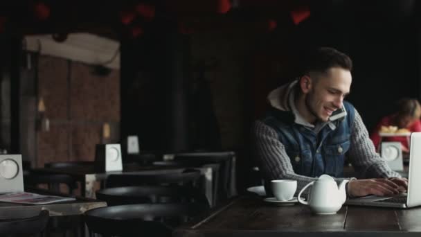 Junge attraktive Mann Multitasking an der halb leeren Café Kerl Antworten auf einen Anruf und Eingabe von Text auf dem Laptop zur gleichen Zeit. - Filmmaterial, Video