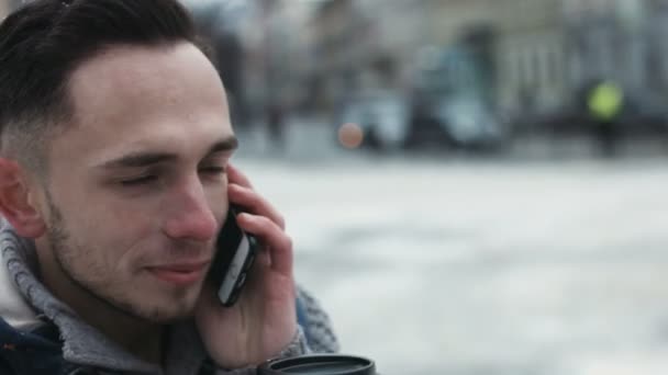 Primo piano di un uomo che parla al cellulare all'aperto il giorno d'inverno. Giovane maschio hanno una dolce conversazione al telefono in attesa di un appuntamento e sorseggiando un caffè per andare
. - Filmati, video