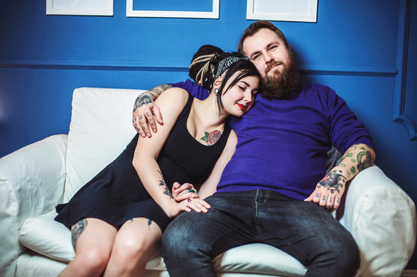 junges verliebtes Paar mit Tätowierungen umarmt und Händchen haltend auf dem Sofa sitzend - Foto, Bild