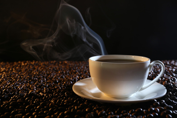 Kop warme koffie onder koffie bonen op donkere achtergrond - Foto, afbeelding