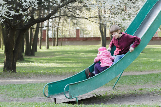 Сидячі діти ковзають вниз на старому парку дитячий майданчик слайд на квітучому весняному фоні фруктового дерева
 - Фото, зображення