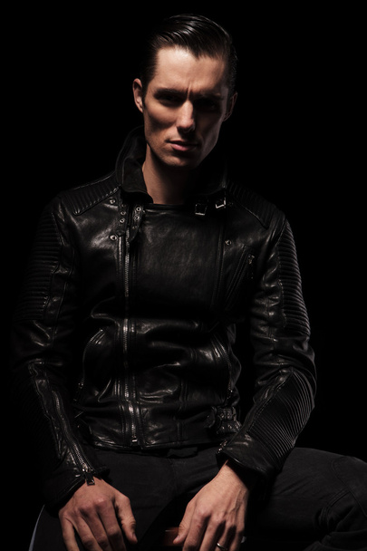 sexy biker in black leather jacket posing seated in dark - Fotoğraf, Görsel