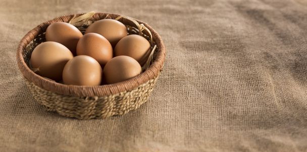 raw organic brown egg in straw basket foreground, on rough fabric - Photo, Image
