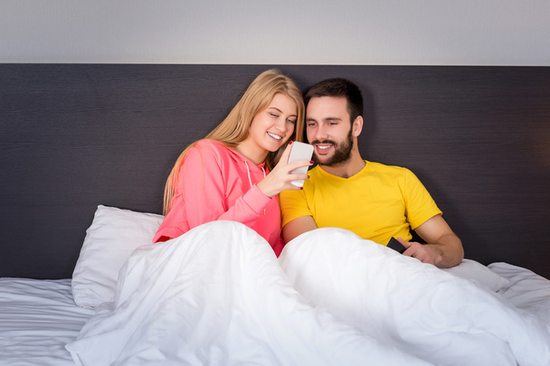 junges süßes Paar im Bett beim Anblick eines Mobiltelefons - Foto, Bild