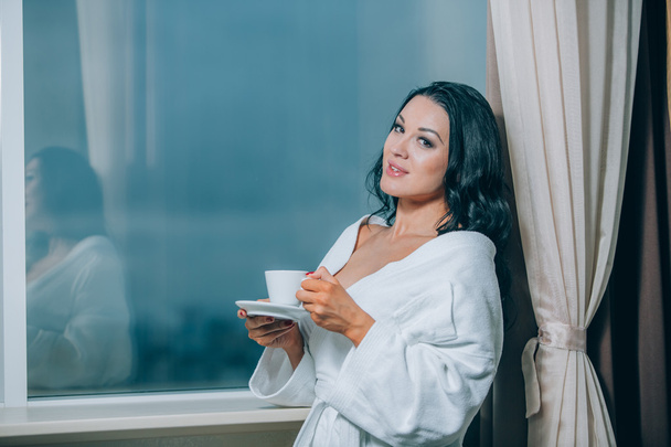 Getting warm with fresh coffee. Beautiful young woman in white bathrobe drinking coffee and looking through a window - Foto, Imagen