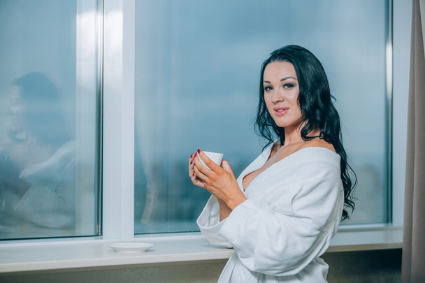 Getting warm with fresh coffee. Beautiful young woman in white bathrobe drinking coffee and looking through a window - Foto, afbeelding