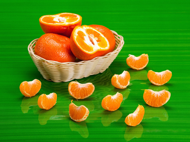 Mandarijnen liggend in een rieten mand voor vruchten, omringd door segmenten voor tangerine, op groene glas. - Foto, afbeelding