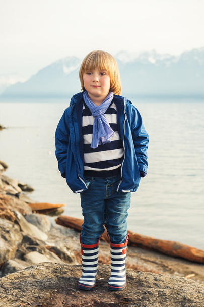 Ritratto all'aperto di adorabile bambino biondo di 4-5 anni, che si diverte in riva al lago in una bella giornata di sole primaverile, indossa una calda giacca blu, sciarpa, jeans denim e stivali da pioggia a righe
 - Foto, immagini