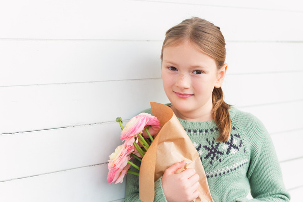Retrato de niña adorable de 8-9 años, vistiendo un jersey verde cálido, sosteniendo flores de color rosa primavera, de pie sobre fondo de madera blanca, concepto del día de la madre
 - Foto, Imagen