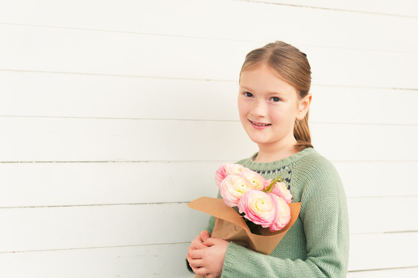 Portrait of adorable little girl of 8-9 years old, wearing warm green pullover, holding spring pink flowers, standing against white wooden background, mother's day concept - Φωτογραφία, εικόνα
