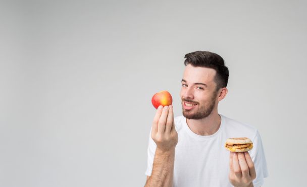 Bearded man in a white shirt on a light background holding a hamburger and an apple - Foto, Bild