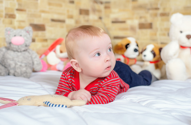 Cute baby boy in jeans and striped t-shirt lying on a bed looking up - Фото, изображение