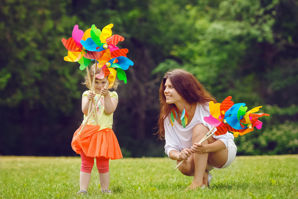Portrait d'une mignonne adorable petite fille en jupe rouge et sa mère ou sa sœur, femmes blanches caucasiennes, tenant un jouet de blanchisseur de moulin à vent dans sa main debout à l'extérieur dans la prairie sur l'herbe verte, concept d'amusement d'été
 - Photo, image