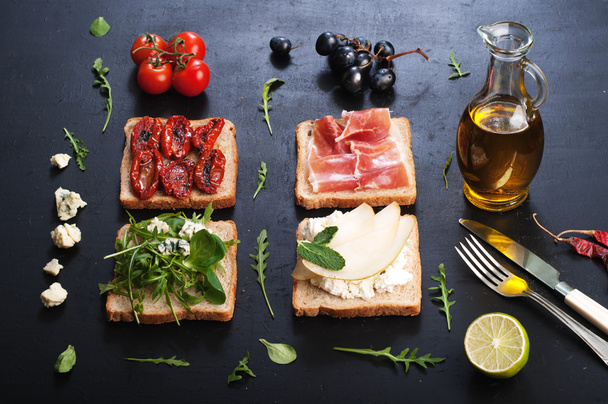 Sandwiches with a variety of toppings on a dark surface. Sandwiches with arugula, leaf mash, blue cheese, sun-dried tomatoes, slices of pear and ham. The concept of useful home cooking - Fotoğraf, Görsel