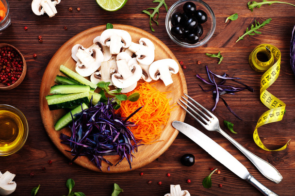 The concept of dietetic vegetarian food. Bright juicy shredded vegetables, such as carrots, purple cabbage, mushrooms and cucumbers, which lies on a circular wooden cutting board. Natural organic products, ready to eat - Фото, зображення