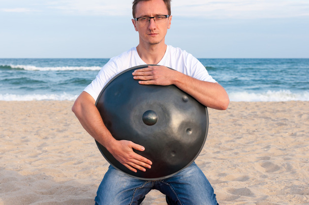Young stylish guy sitting on the sand beach and holding a handpan or hang with sea On Background. The Hang is traditional ethnic drum musical instrument - Fotografie, Obrázek