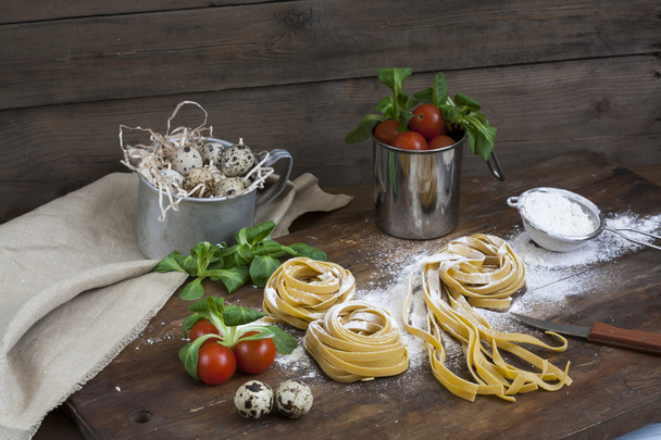 Raaka kotitekoinen pasta, viiriäisen pääsiäismunia alumiinimukiin, vihreää salaattia, tomaatteja ja jauhoja puupöydällä
 - Valokuva, kuva