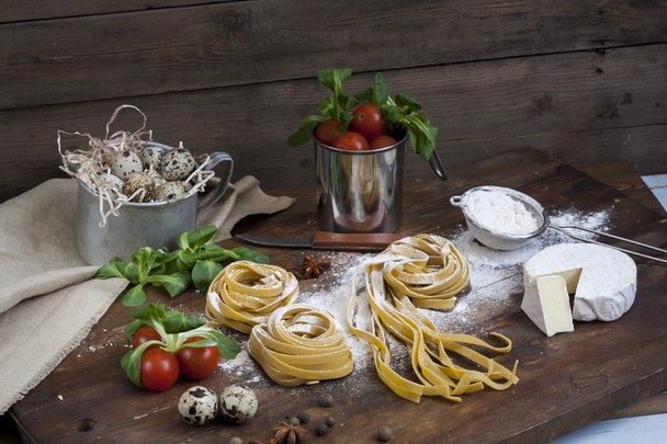 Raw homemade pasta, quail easter eggs in a aluminum cup, green lettuce, tomatoes and flour on wooden table - Foto, Imagen