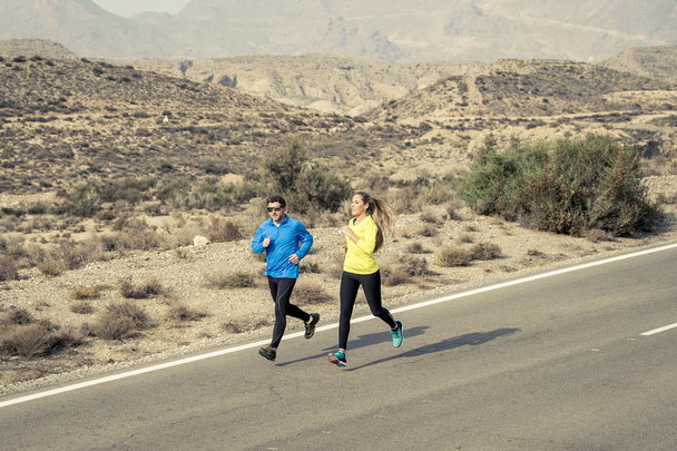 attractive sport couple man and woman running together on desert asphalt road mountain landscape - Foto, afbeelding