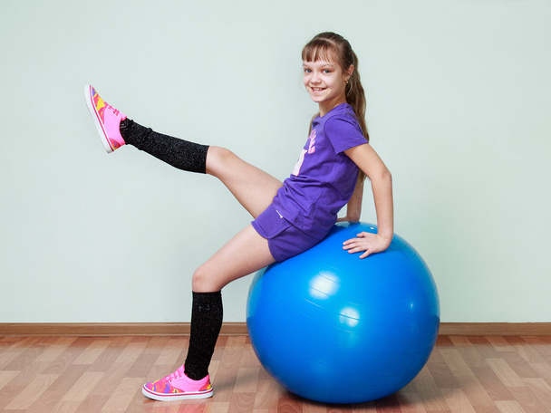 Menina está sentada em uma bola azul em forma, levantando a perna, smi
 - Foto, Imagem