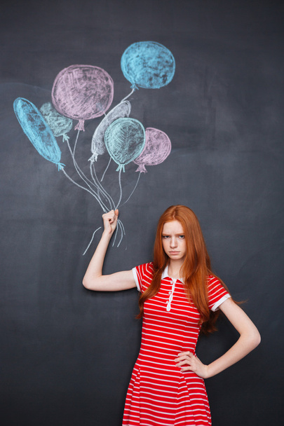 Sad girl holding drawn colorful balloons over blackboard background - Фото, зображення