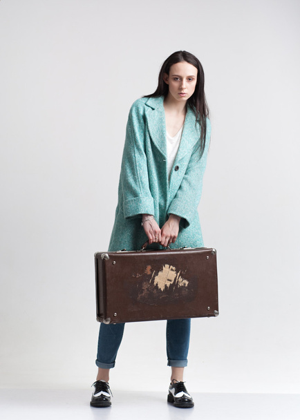 Young fashionable girl in the cyan coat posing with suitcase. Isolated on a grey background. - Фото, зображення