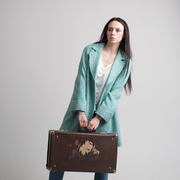 Young fashionable girl in the cyan coat posing with suitcase. Isolated on a grey background. - 写真・画像