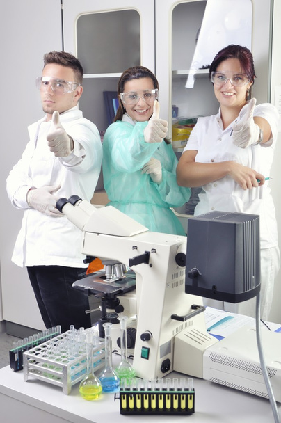 Trabajo en equipo exitoso dentro del laboratorio. Científicos sonrientes mirando la cámara haciendo gestos con los pulgares hacia arriba
. - Foto, Imagen