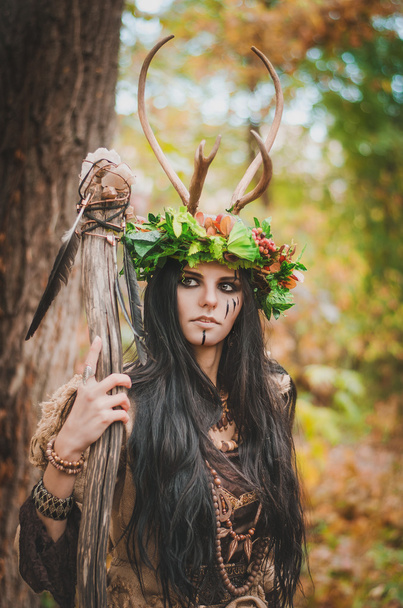 beautiful brunette with painted face, in a flower wreath and a wooden staff in his hand, looking forward belligerently - Zdjęcie, obraz