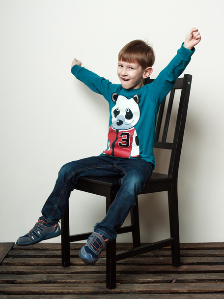  Pieni hauska poika istuu tuolilla kädet ylhäällä hymyillen.
 - Valokuva, kuva