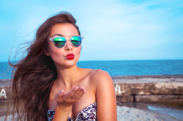 Outdoor summer portrait of young pretty woman looking to the ocean and sending kisses, enjoy her freedom, wearing stylish sunglasses and clothes. - 写真・画像