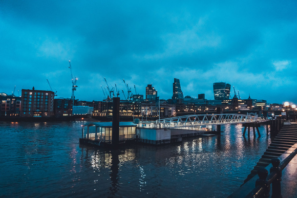 Deserted Ferry Dock Illuminated at Night on Thames River with City Skyline and Construction Cranes in Background with Cloudy Evening Sky - Photo, Image