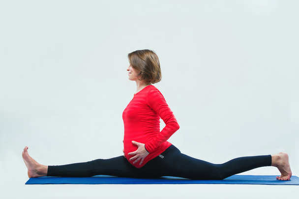 Mujer embarazada practicando yoga, foto de estudio, sentado en las divisiones, las manos sosteniendo el abdomen
 - Foto, imagen