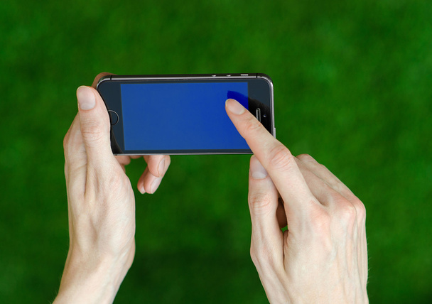 Human hand holding a modern mobile phone with a blue screen on a background of green grass first-person view - Fotografie, Obrázek